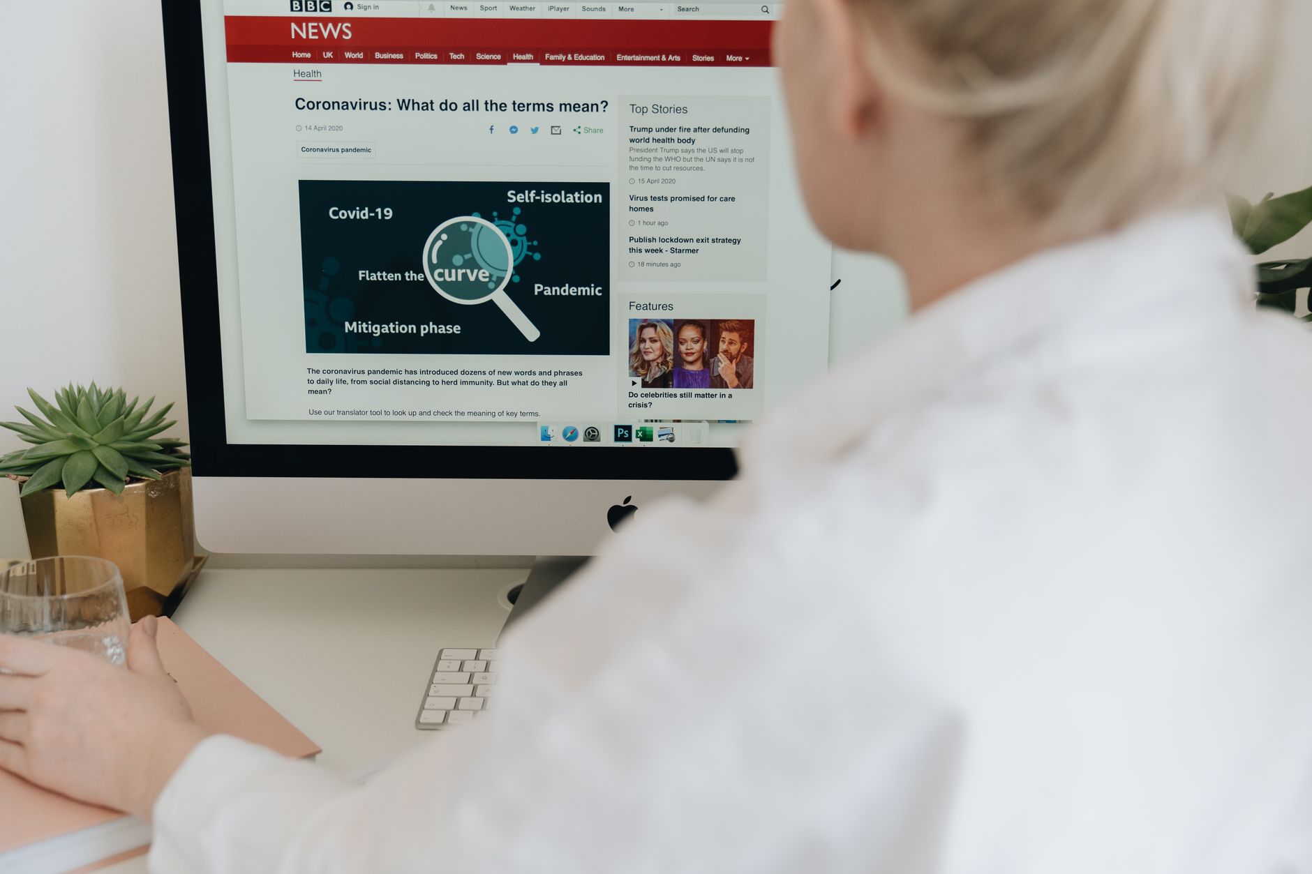 A person at a desk compares a video on a phone with a news website open on a laptop, index finger pointing at the screen.
