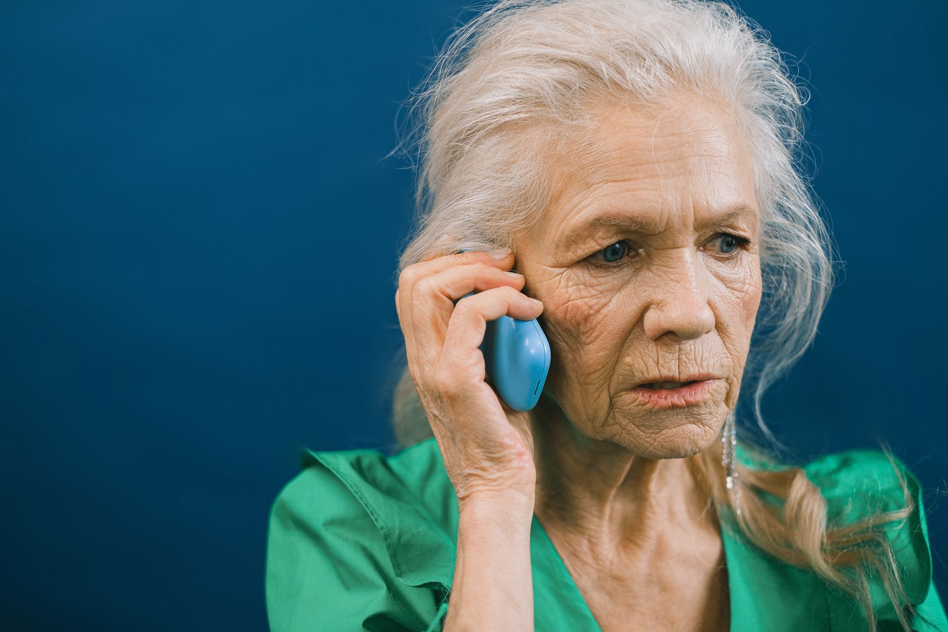 An older adult sits on a couch answering a landline phone with a worried expression in a dim living room.