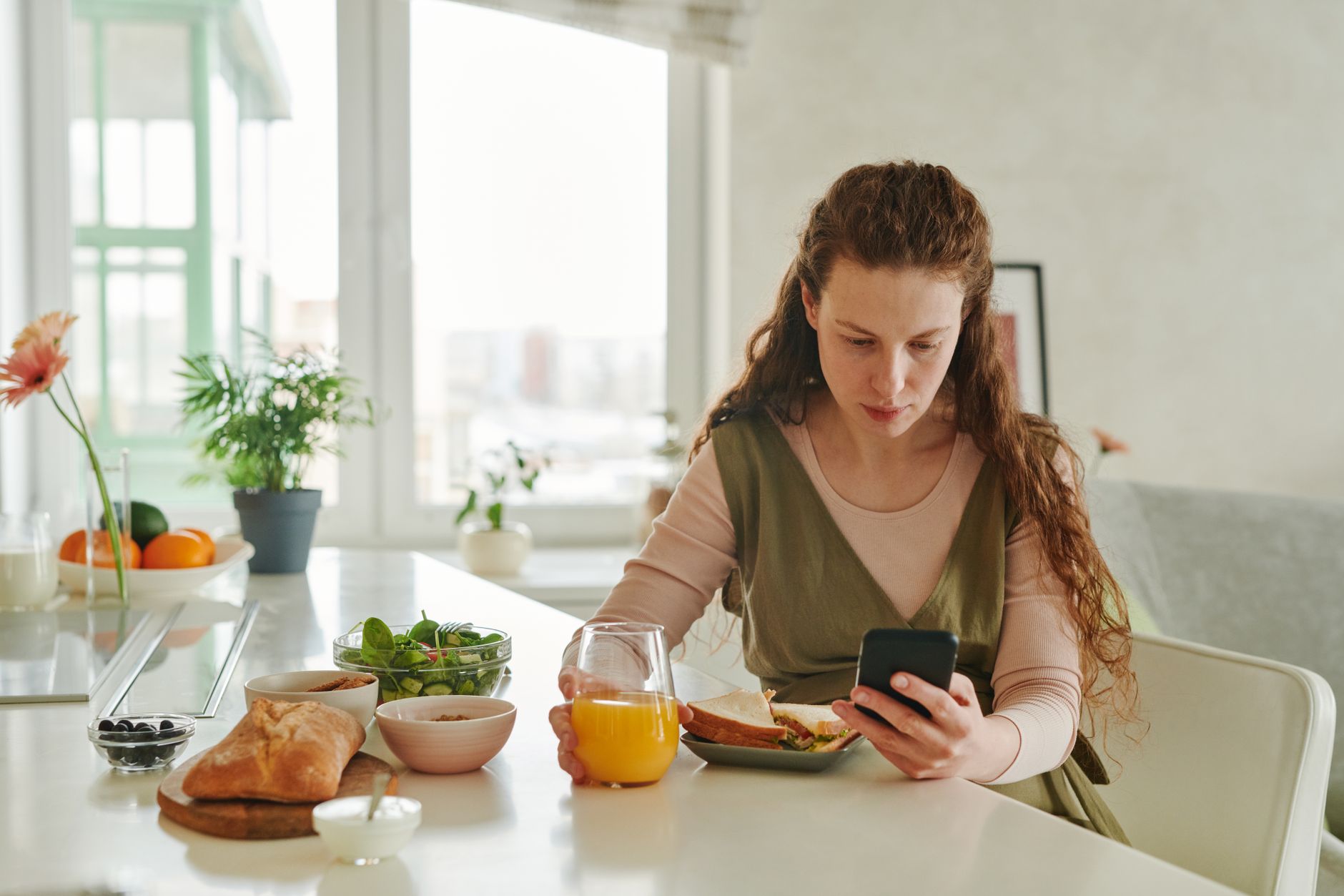 An adult at a kitchen table holds a smartphone with a concerned expression while looking at a notepad with family phone numbers.