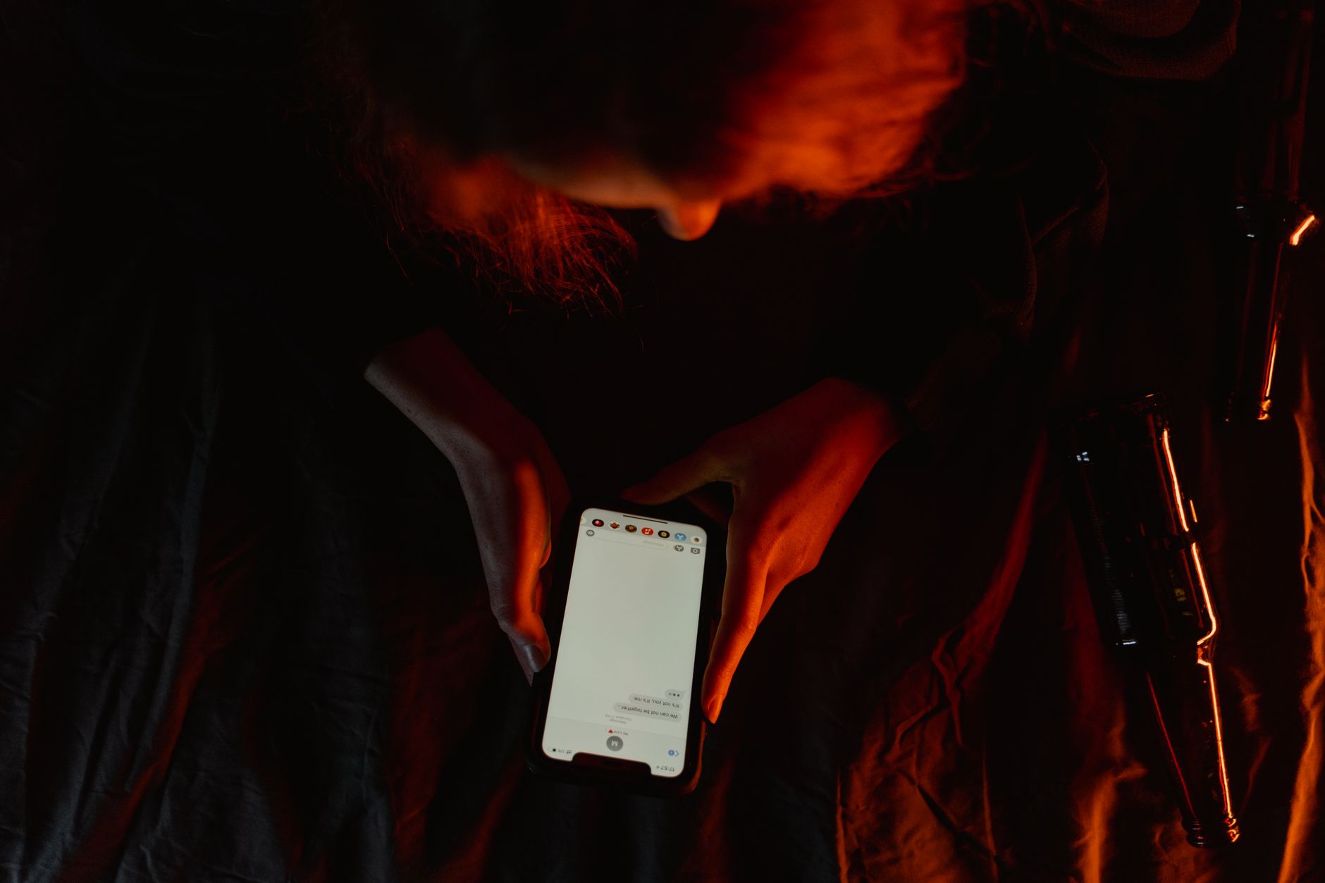 A person sits alone in a dark living room holding a smartphone and looking worried while messaging.